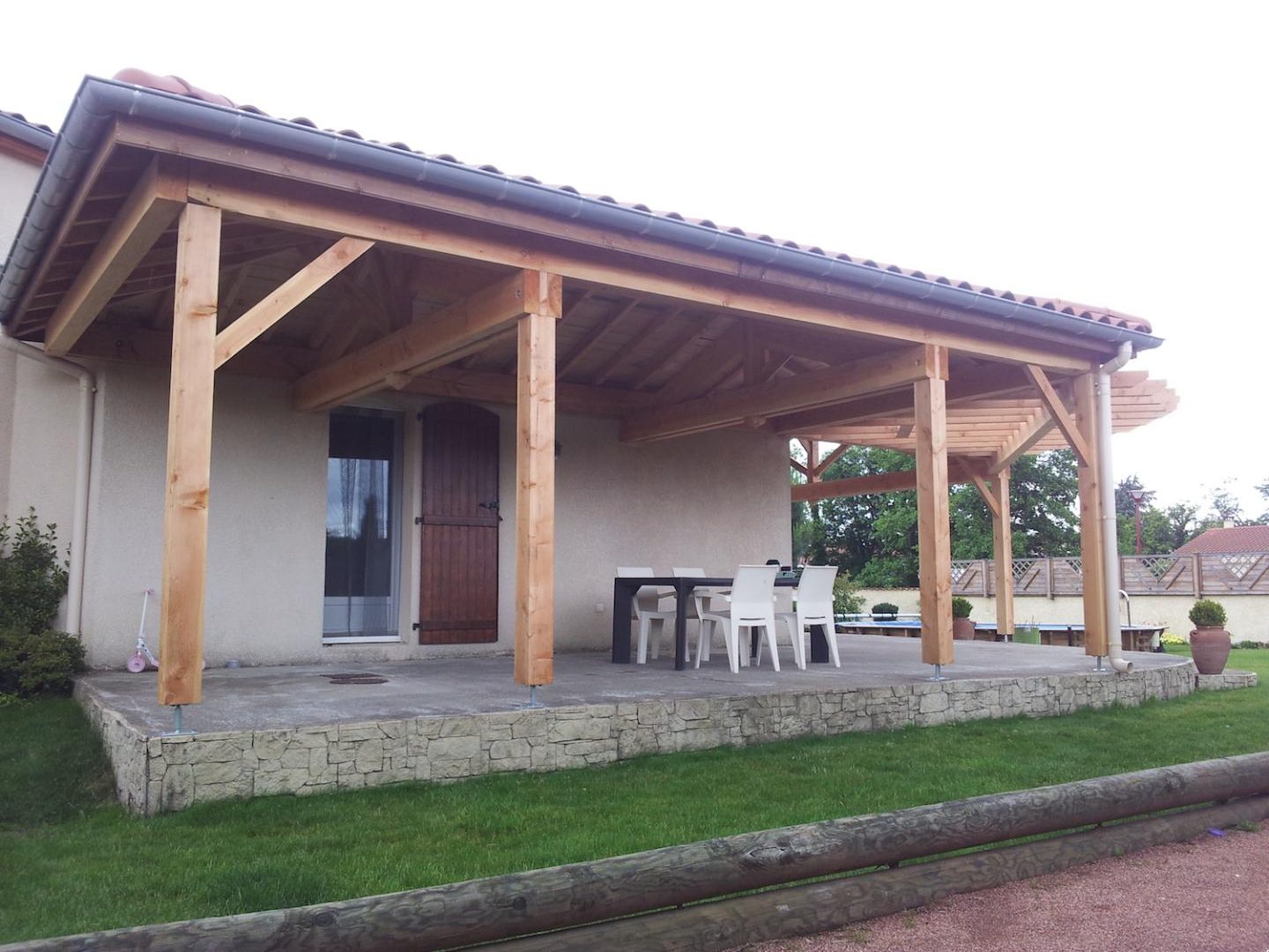 Préau en bois couvert de tuiles adossé à une maison, au-dessus d’une terrasse